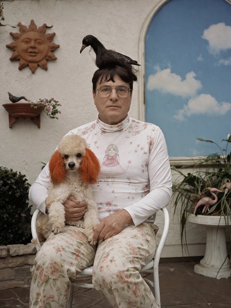 A slightly surreal but photorealistic portrait of a seated person in a quiet courtyard or patio, centered in frame, wearing a fitted white long-sleeve turtleneck top with delicate pink fl...
