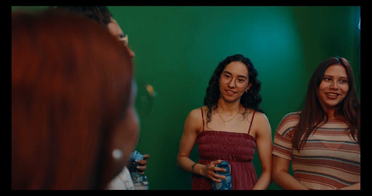 A cinematic candid photo of 4 young adults standing in front of a smooth studio green screen wall, framed like a behind-the-scenes chroma key test. In the extreme left foreground, a woman...