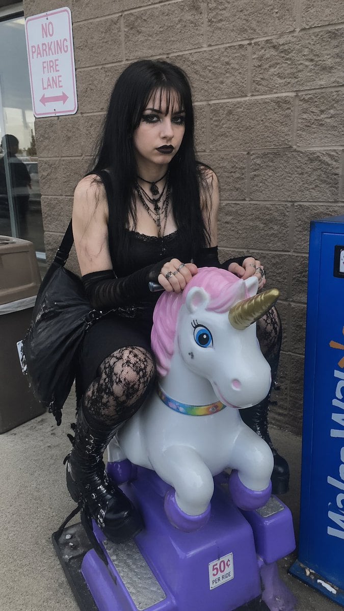 Generates a realistic, candid photo of a goth girl sitting on a children's coin-operated ride outside a store.