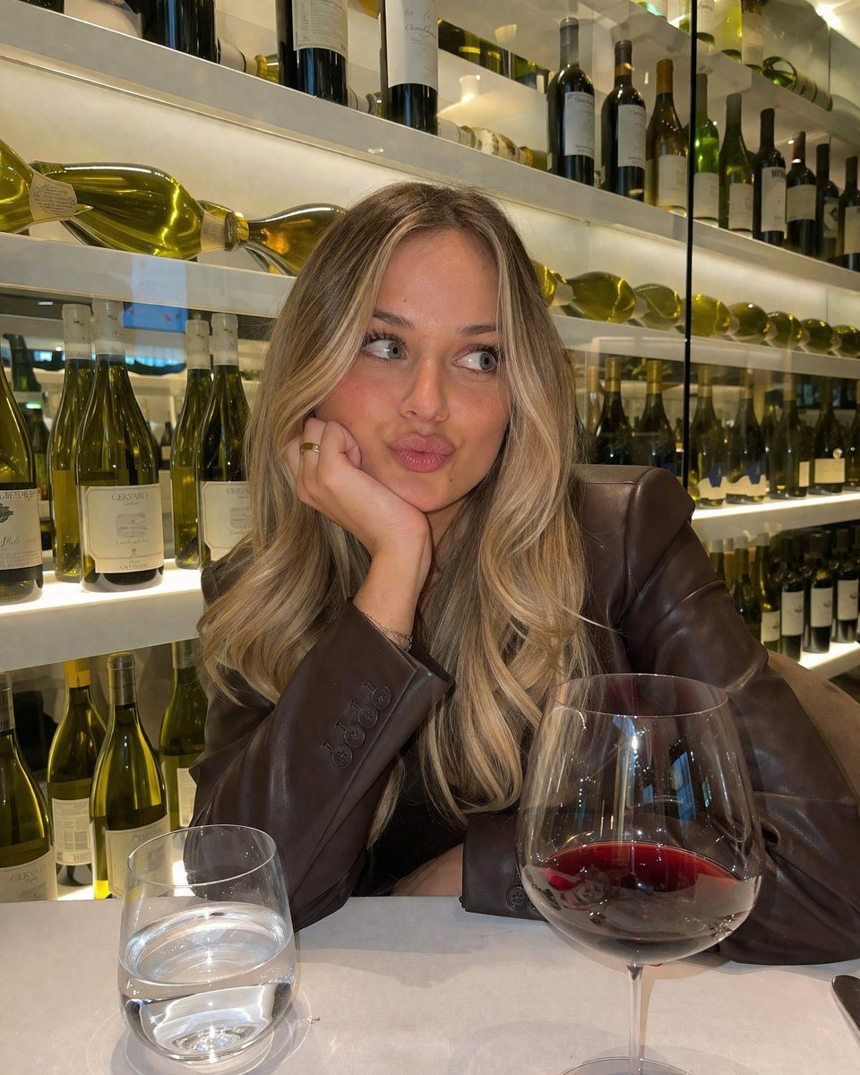 A photorealistic, casual smartphone photo of a young woman with long blonde balayage hair sitting at a table in a wine bar. She is wearing a dark brown leather blazer. She is resting her...