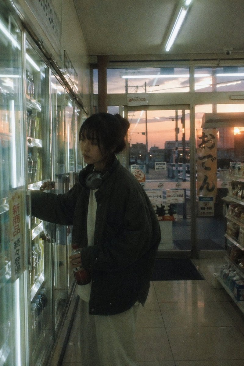 Japanese negative film aesthetic, convenience store scene at dusk, mixed cool indoor light and warm outside light, slight highlight bloom, low contrast, muted colors, subtle grain, subjec...