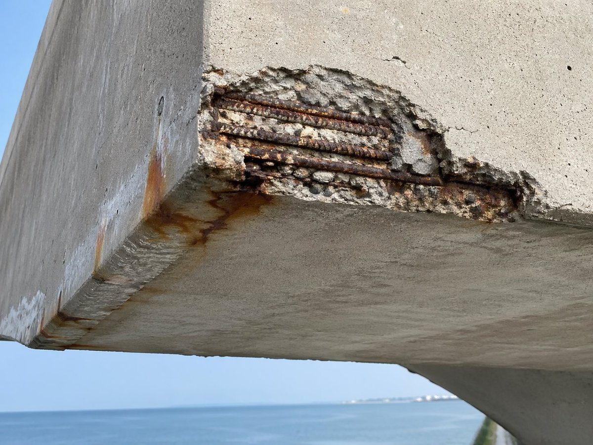 A hyper-realistic close-up documentary photograph of severe chloride-induced corrosion damage on a reinforced concrete coastal structure, viewed from slightly below at the corner of a can...