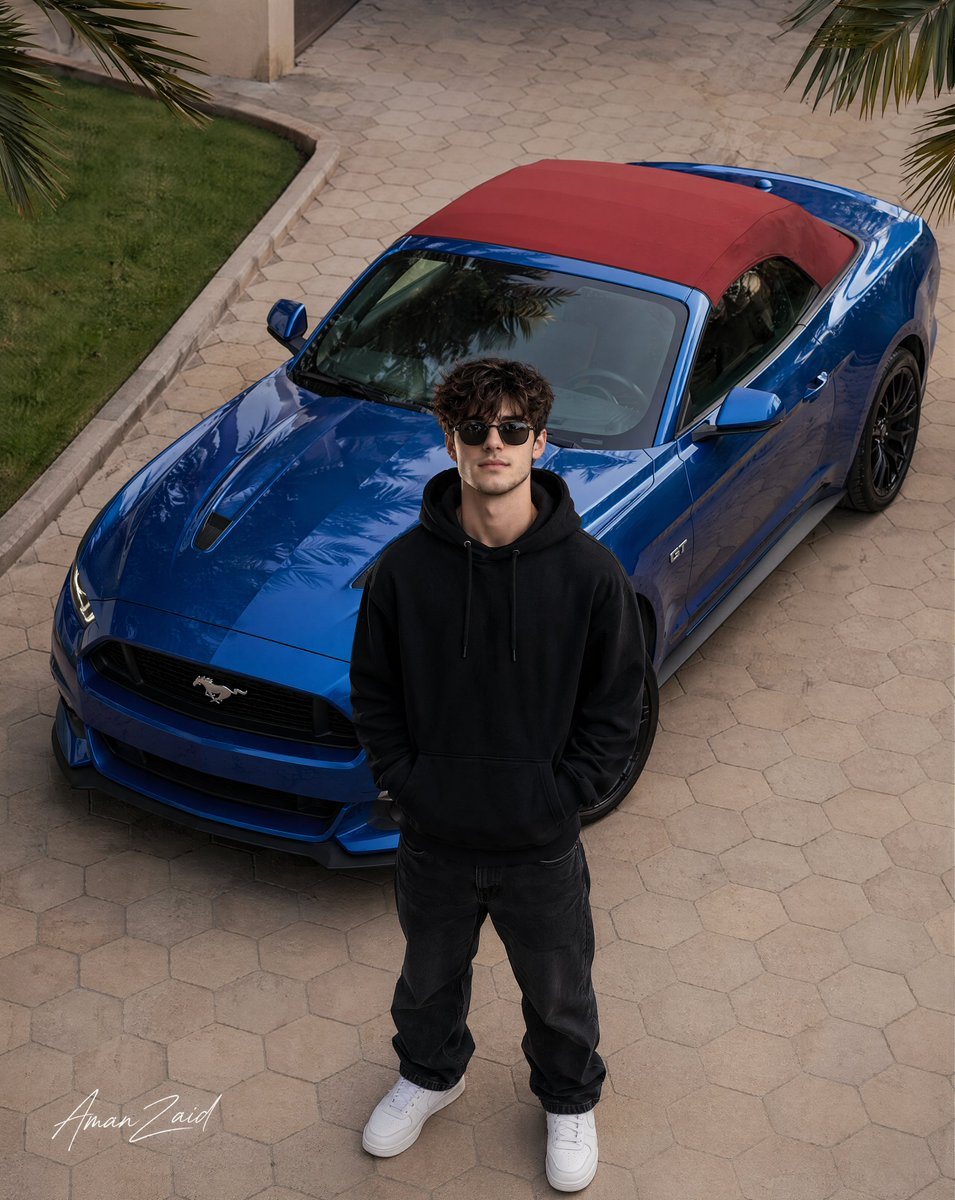 一份全面的超写实提示词,用于拍摄一位时尚年轻男子与定制版 Ford Mustang 合影的高角度航拍照片,包含关于面部准确性、衣物纹理和光影的详细说明。