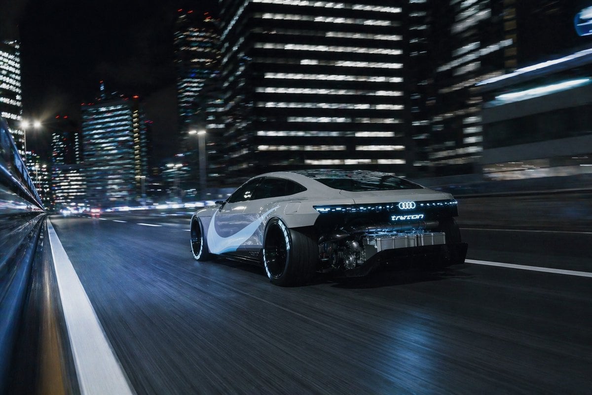 A cinematic nighttime automotive action shot of a futuristic electric sports coupe racing through a modern downtown city, viewed from a low rear three-quarter tracking angle. The car is a...