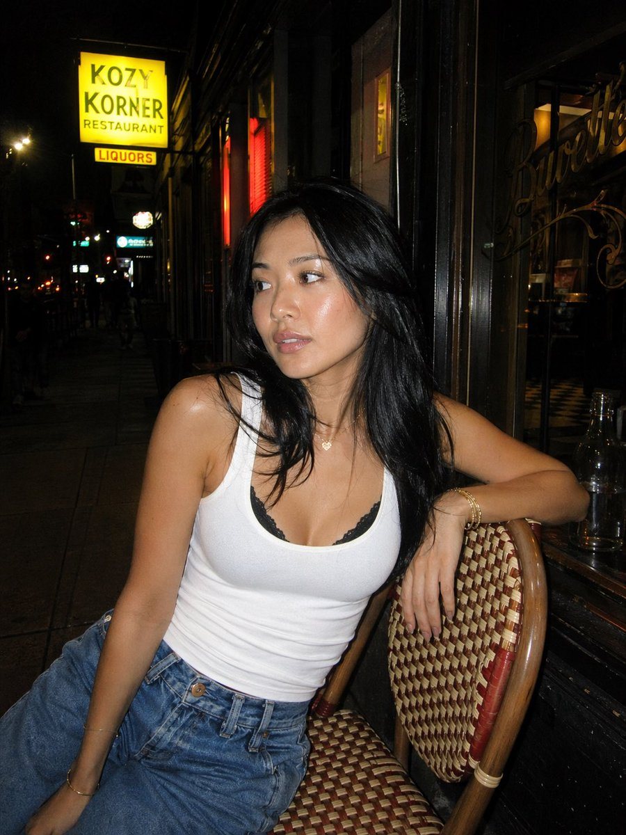 A candid nighttime street portrait of a young woman sitting casually on a woven bistro café chair outside a small urban restaurant. She has long black hair worn loose and slightly tousled...