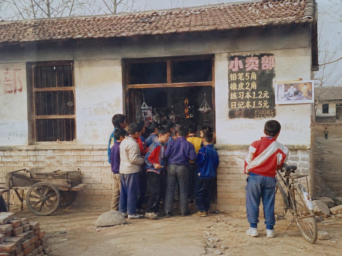 A highly realistic documentary-style photo of a 1998 rural elementary school gate-side convenience stall in northern China, shot like an ordinary family snapshot on slightly faded film. T...