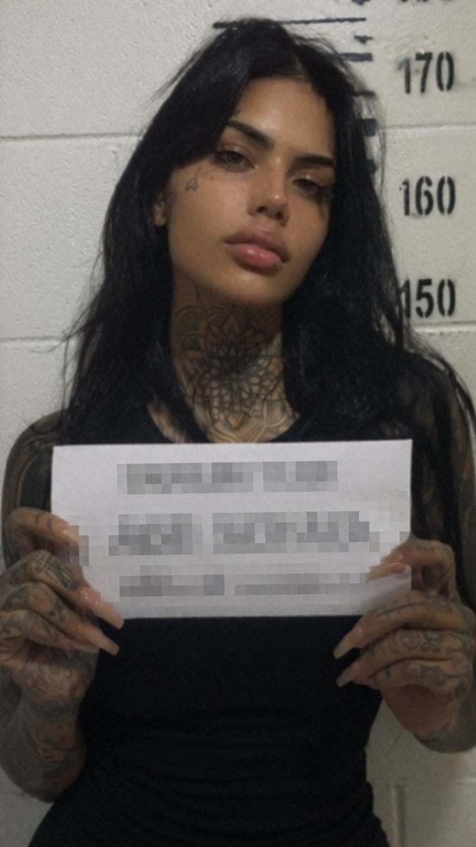 A realistic, slightly grainy, low-lighting mugshot of a young woman with long, messy black hair, prominent cheekbones, and full lips. She has a small tattoo near her right eye and extensi...
