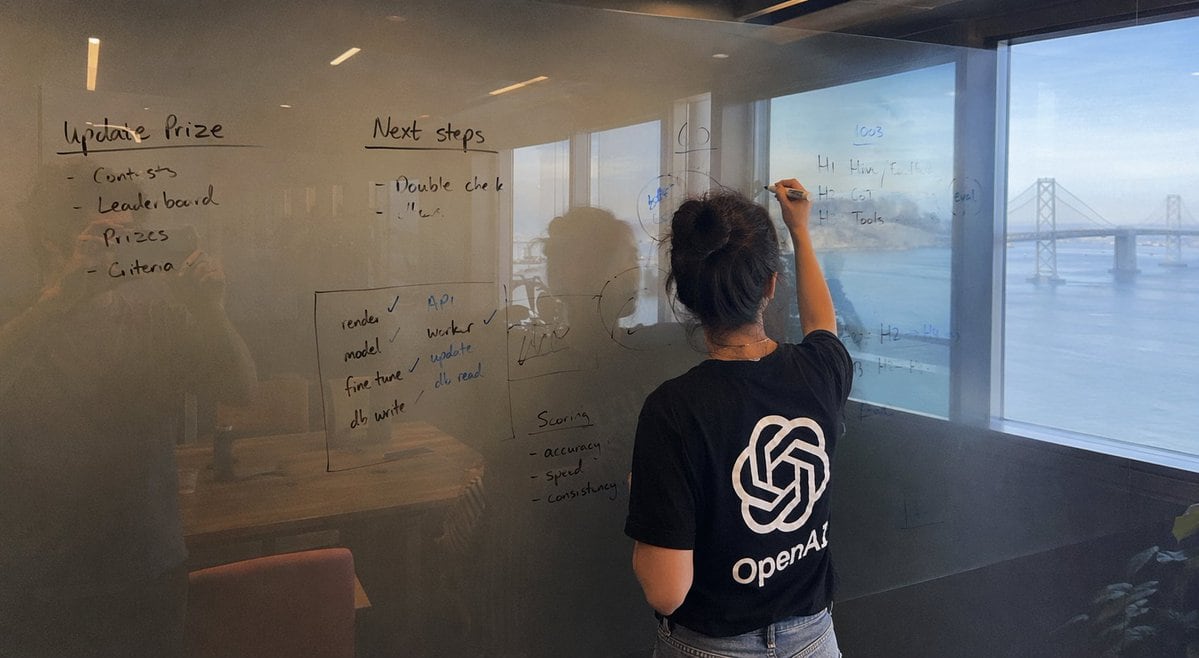 A wide image shot on iPhone of a glass whiteboard, in a room overlooking the Bay Bridge. The field of view shows a woman writing, sporting a tshirt with a large OpenAl logo. The handwriti...