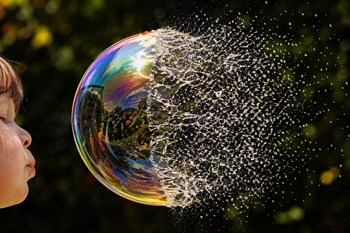 A high-speed macro photograph of a soap bubble bursting in a sunlit garden, framed horizontally. On the far left edge, show the side profile of a young child with visible cheek, nose, lip...