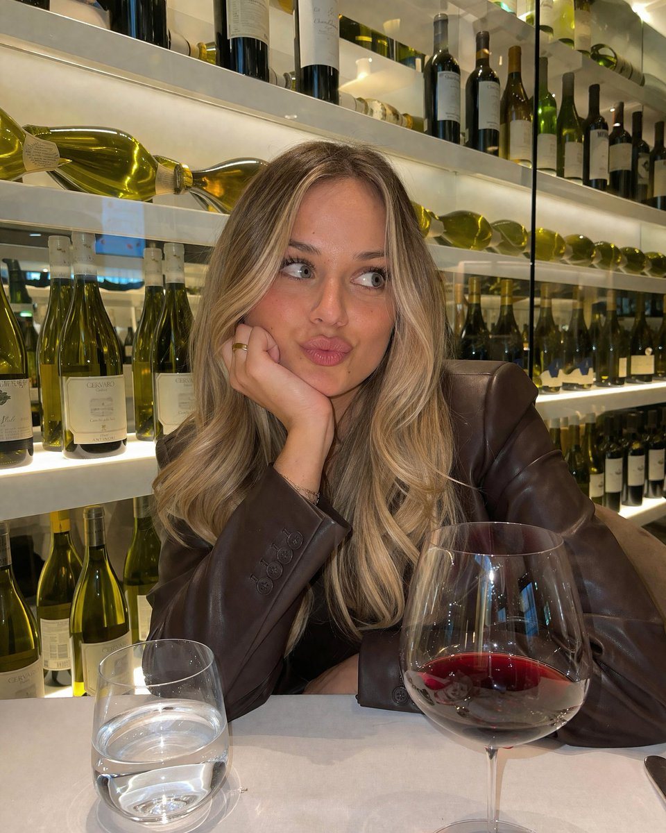 A candid, photorealistic portrait of a young woman with long blonde hair wearing a dark brown leather blazer. She is sitting at a table in a restaurant or wine bar, resting her chin on he...