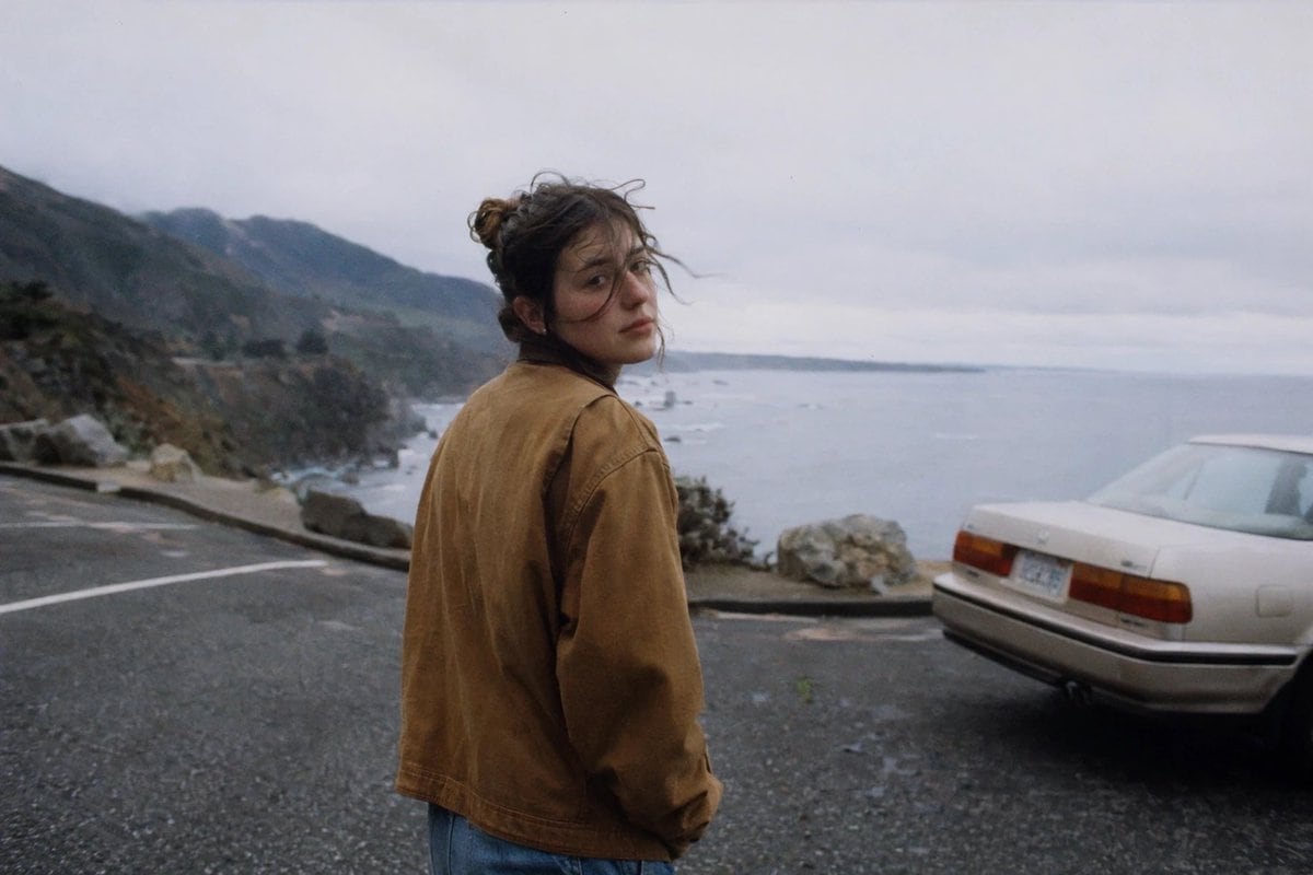 A cinematic candid photo of a young woman standing in a wet coastal roadside turnout on an overcast day, viewed from behind and slightly to the side as she turns her head back toward the...