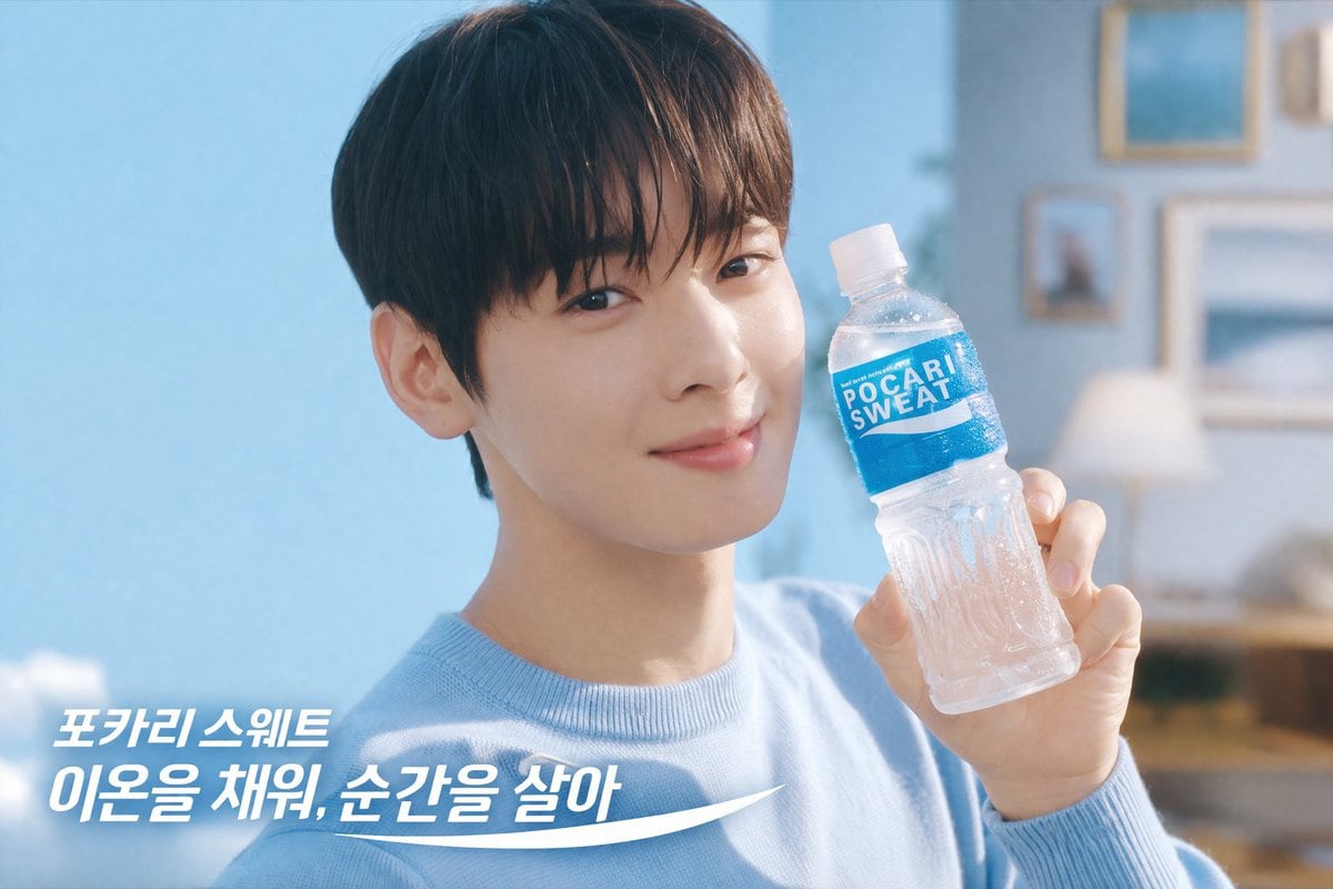 A bright, refreshing commercial photography shot of a young handsome East Asian man with dark bangs wearing a light blue crewneck sweater. He is smiling gently at the camera and holding u...