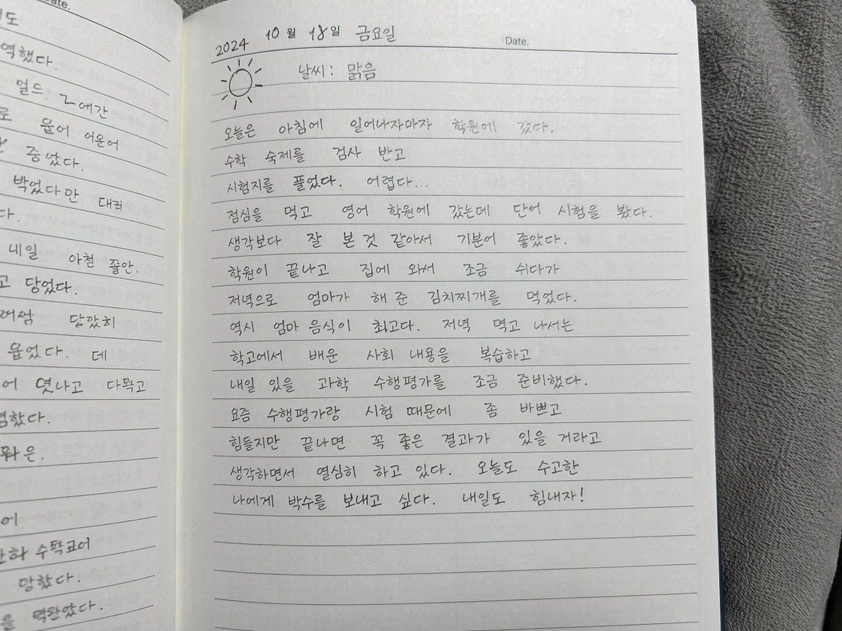 A photorealistic, casual top-down snapshot of an open lined notebook resting on a textured gray fabric surface. The notebook pages are filled with neat, legible Korean handwriting. The ri...