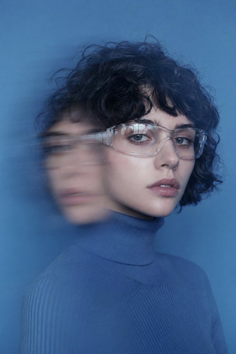 portrait of a young woman with short curly black hair and fair skin, wearing transparent safety goggles and a blue ribbed turtleneck sweater, seated in front of a solid azure blue backgro...