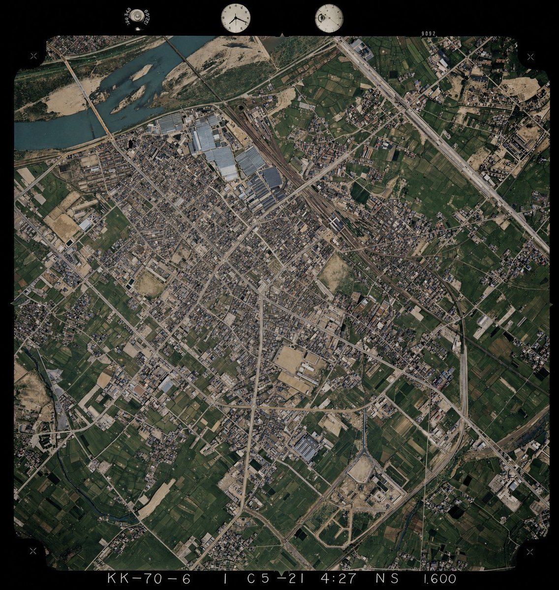 Using REFERENCE_0 as the base image, colorize the monochrome historical aerial photograph while preserving the exact original framing, angle, scale, film border, edge markings, and all ge...