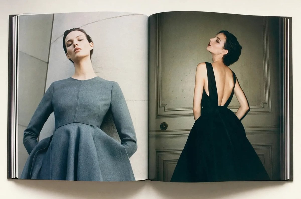 A high-end editorial fashion photograph shown as an open large-format hardcover magazine or art book lying flat on a clean off-white surface, viewed from directly above. The two-page spre...
