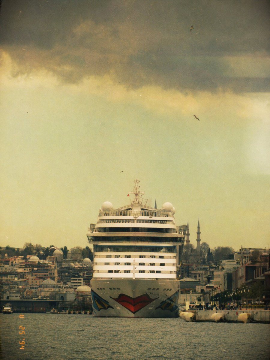 A front-facing photograph of a massive white cruise ship centered in the frame on the water in Istanbul, seen from a low distance across a narrow harbor channel, with the ship moving slow...