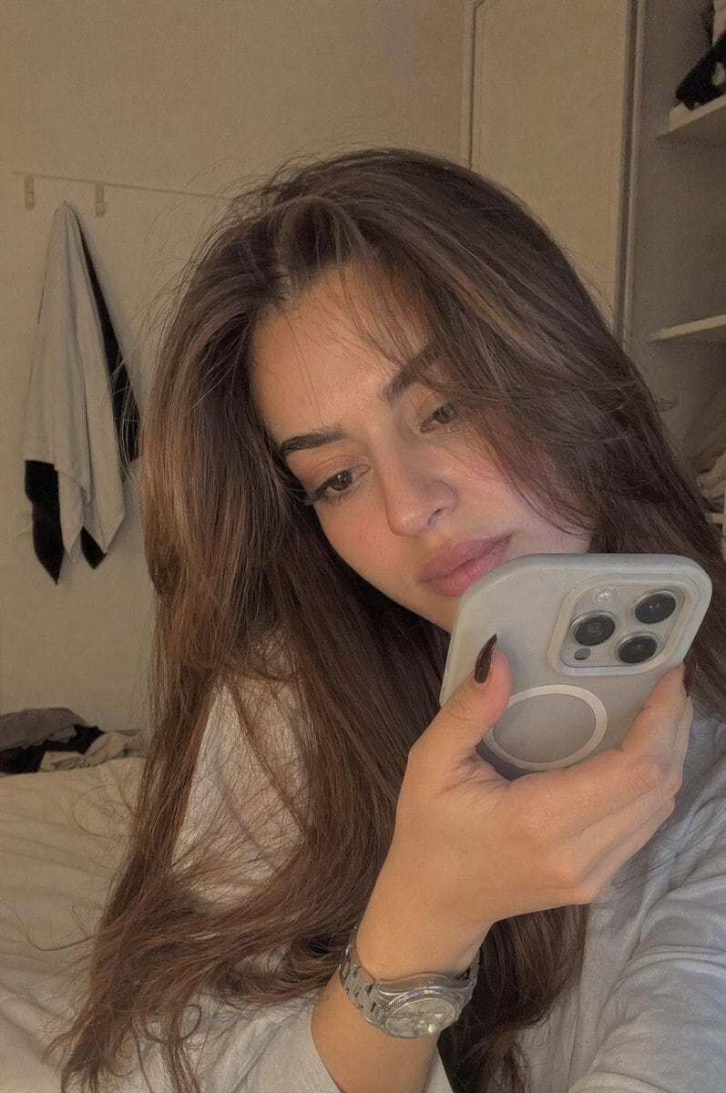 A realistic young woman sitting casually in a softly lit bedroom during late afternoon. She is holding her phone very close to her face as if recording a private video or voice note. Fram...