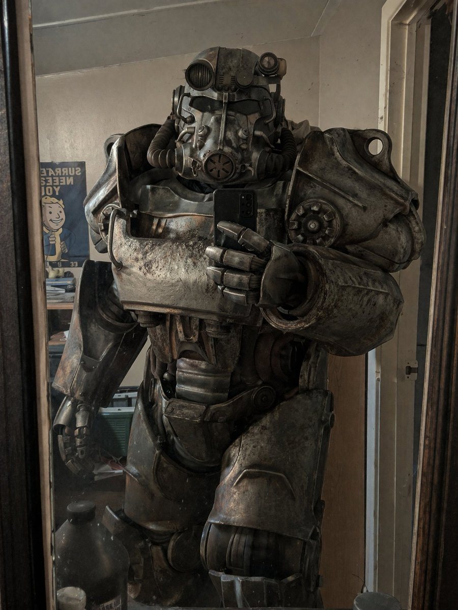 A person wearing a highly detailed, weathered, and battle-damaged T-60 power armor from Fallout taking a mirror selfie with a black smartphone. The armor is bulky, metallic, and covered i...