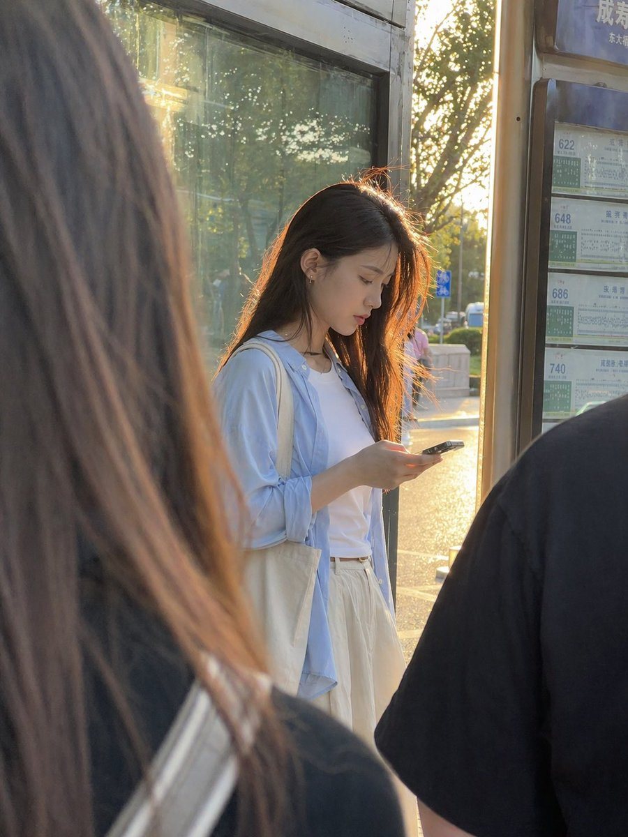 A candid, ultra-realistic street photo of a young East Asian woman standing at a city bus stop during golden hour, viewed from slightly behind other waiting passengers so the composition...