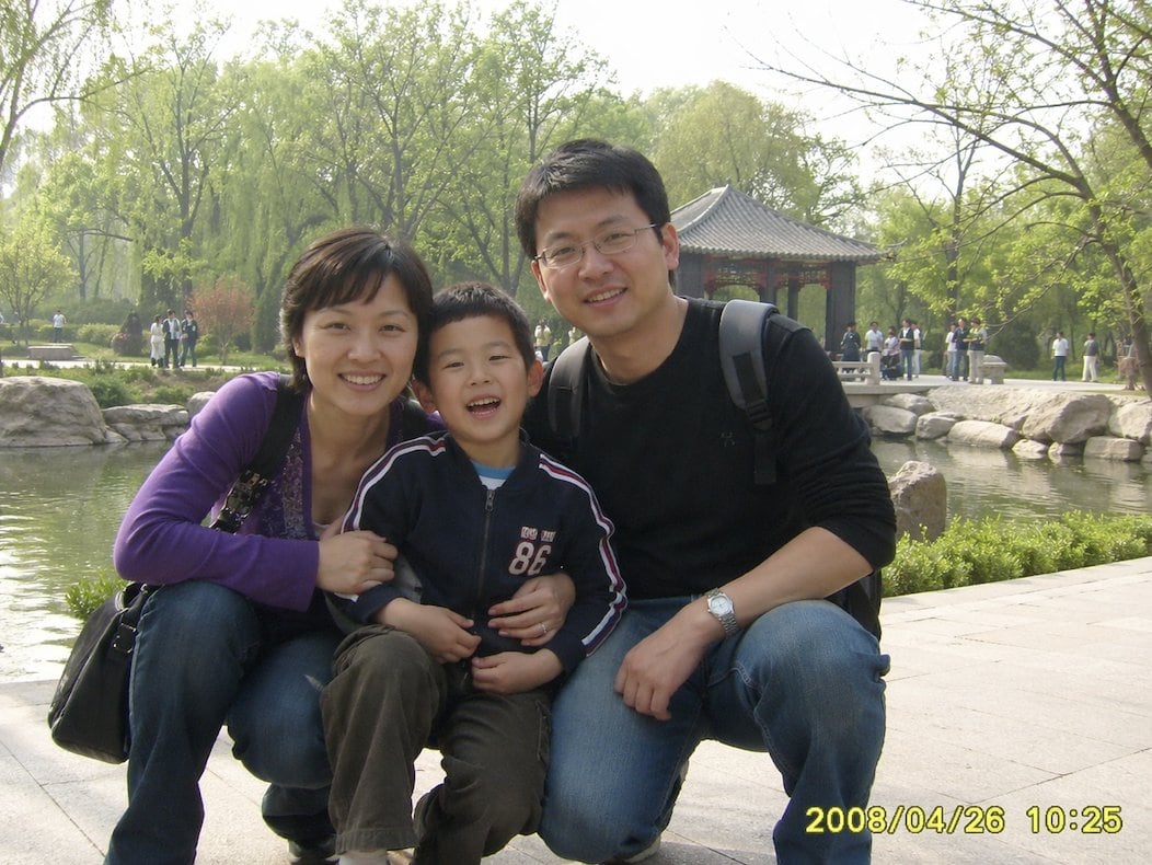 A candid family snapshot styled like a consumer digital camera photo from around 2008, showing a Chinese family of three posing closely together in a public park in springtime. The mother...
