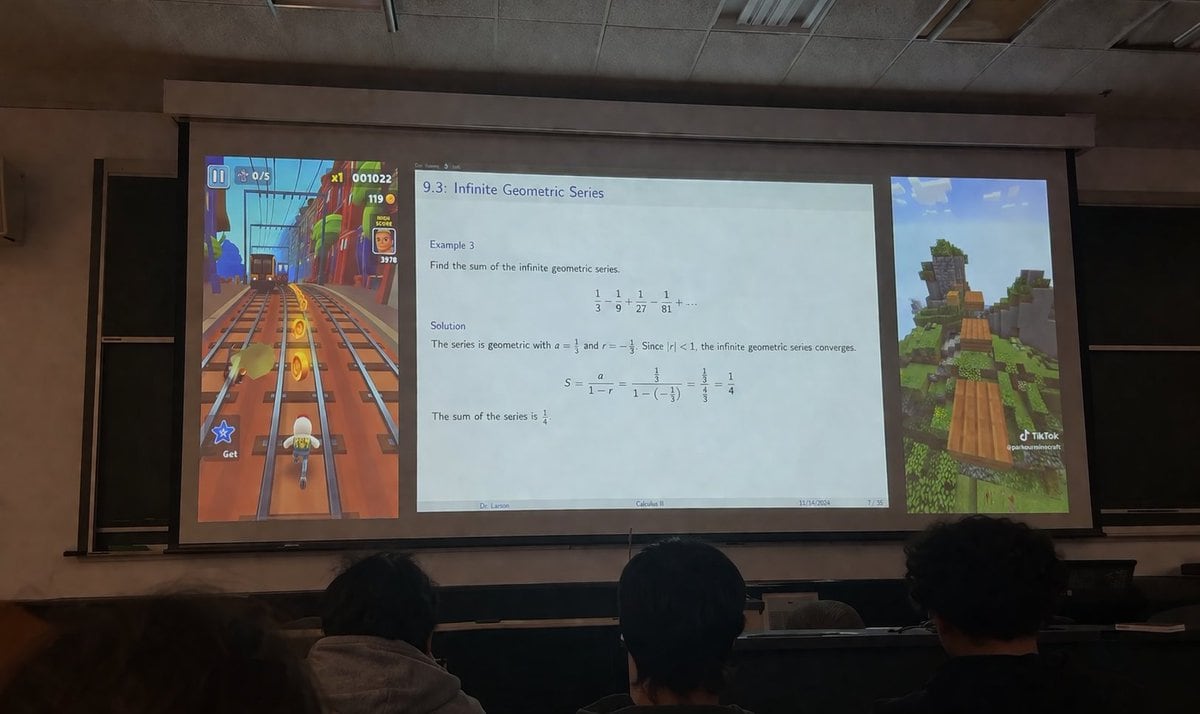 A realistic photograph taken from the audience in a dimly lit university lecture hall, looking over the backs of the heads of three students in the foreground towards a large projector sc...