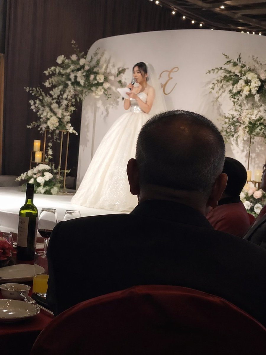 iPhone candid shot, the bride speaking on stage at a wedding, with the father’s back visible as he sits at a banquet table below.