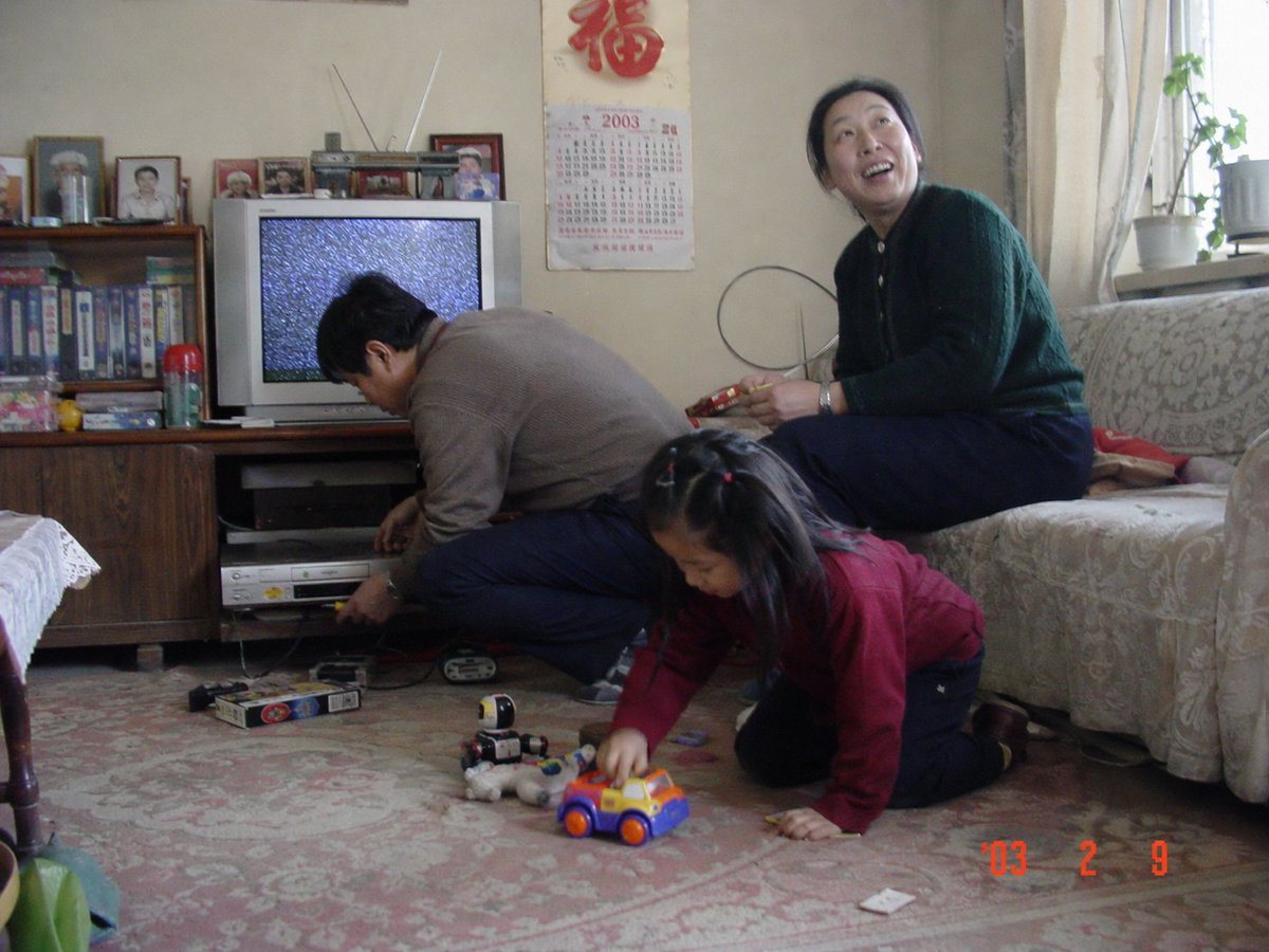 A candid, unprocessed indoor photograph taken with an early 2000s consumer digital camera or cheap film camera. The image features a cheerful Chinese family of three in their modest Beiji...
