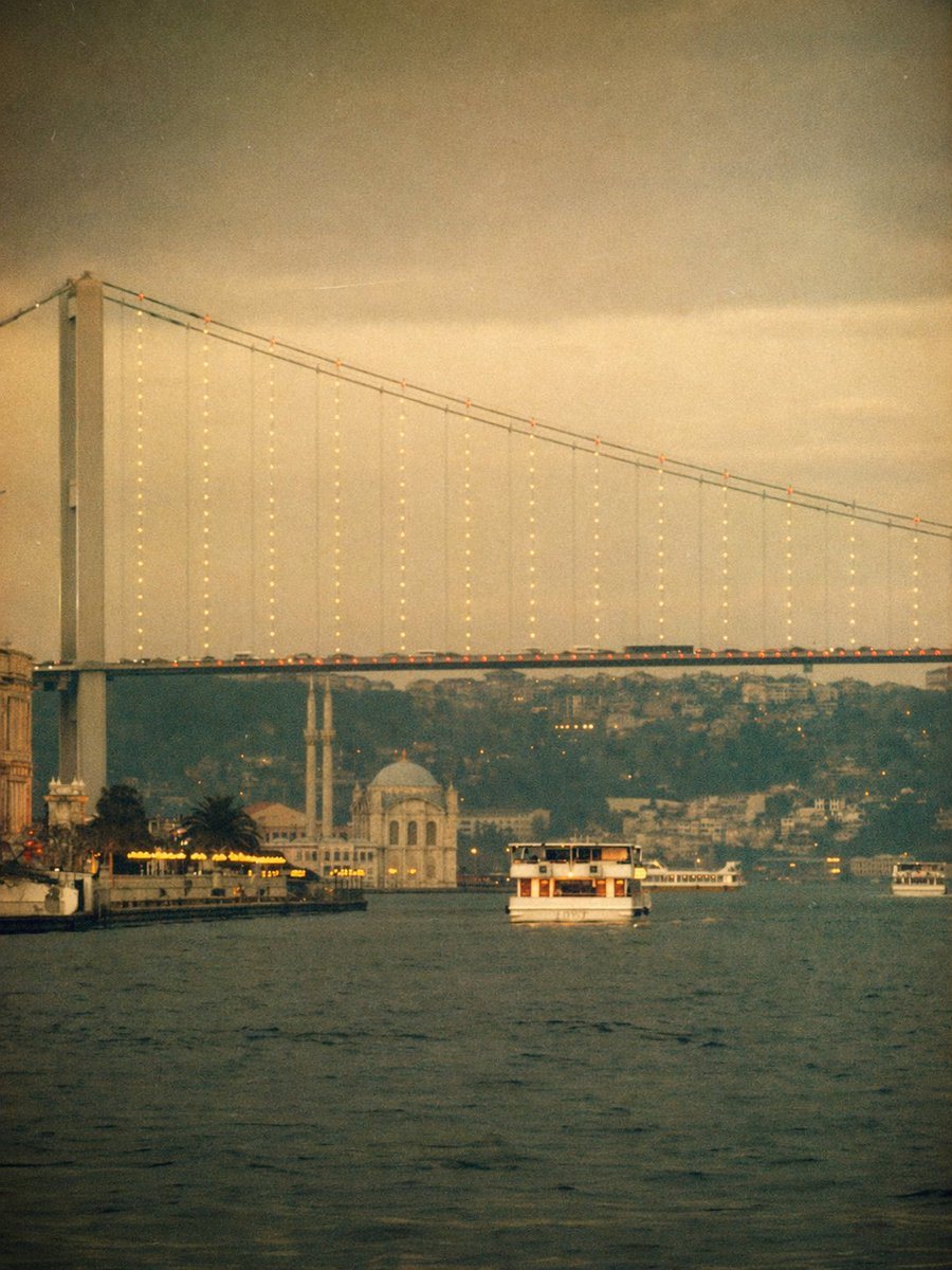 A dreamy vintage-style photograph of the Bosphorus in Istanbul at dusk, shot from water level in a vertical composition. The enormous suspension bridge spans the frame from upper left to...