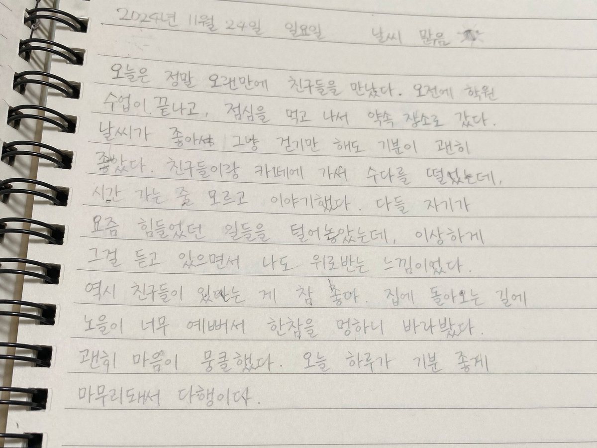 A close-up, slightly angled photograph of a spiral-bound notebook page filled with handwritten Korean text in pencil. The notebook has horizontal ruled lines and a black metal spiral bind...