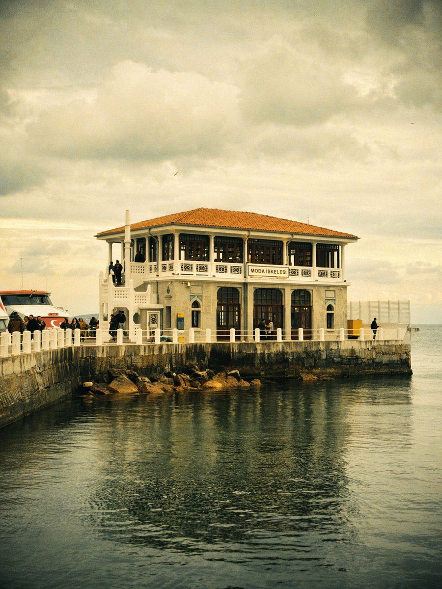 A dreamy vintage travel photograph of the historic Moda pier terminal in Istanbul, centered at the end of a stone waterfront jetty, shot in a vertical composition from across calm dark wa...