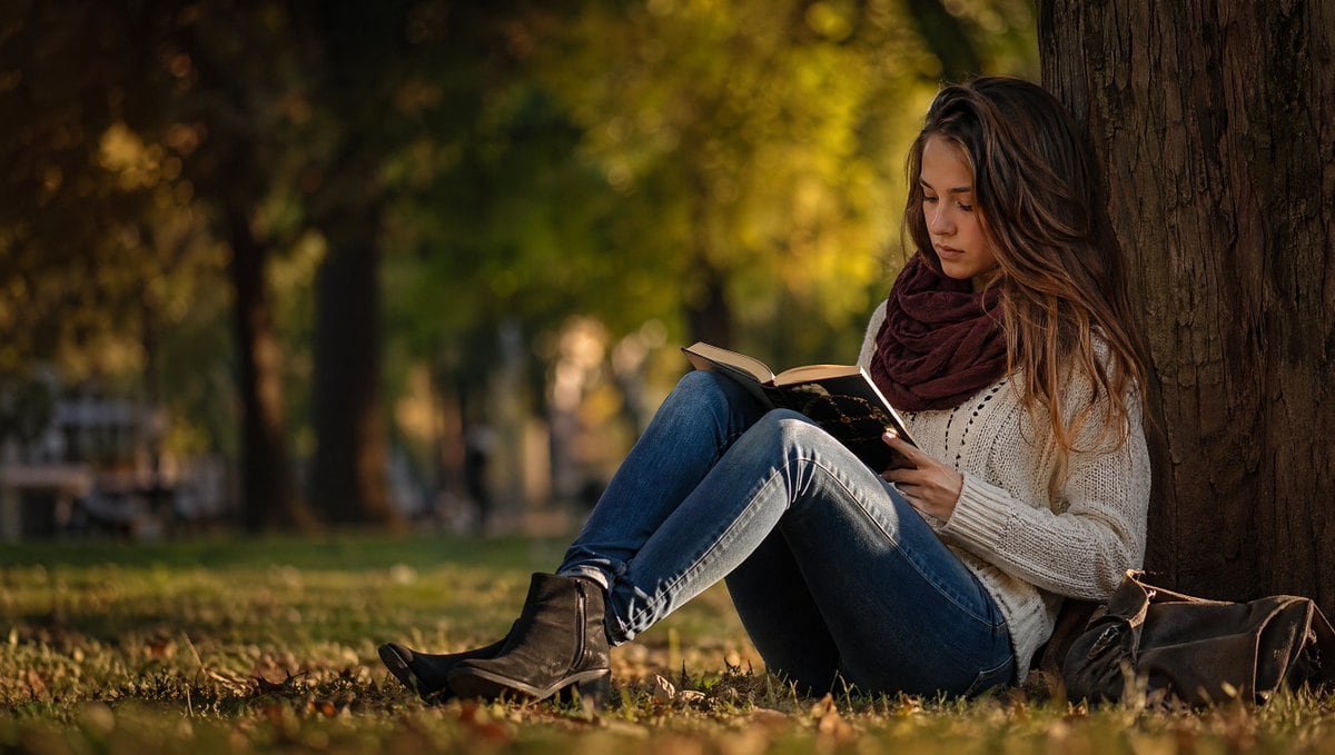 A realistic lifestyle stock photo of a young woman sitting on the ground and leaning against a large tree trunk in a park during golden hour in autumn. She is positioned on the right side...