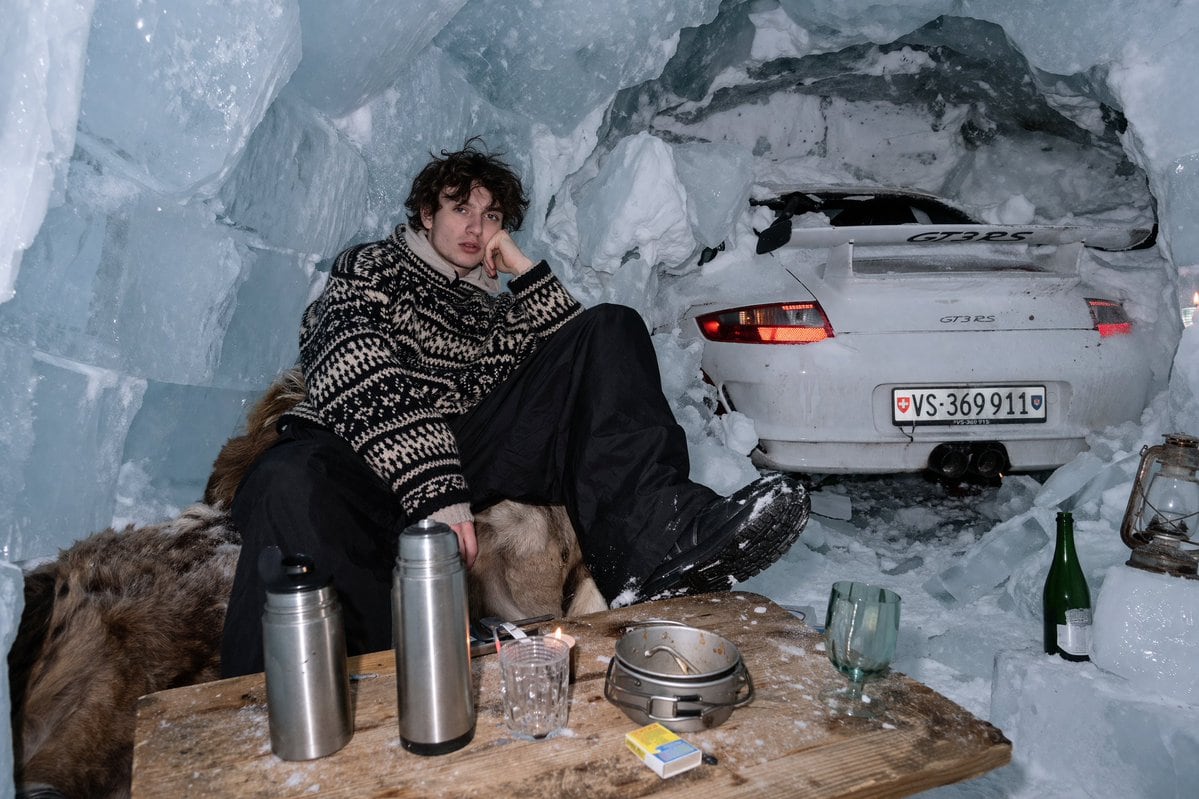 A realistic image shot with harsh direct flash photography inside an ice cave made of large ice blocks and snow. A young man with messy dark curly hair sits on a brown fur pelt, leaning h...