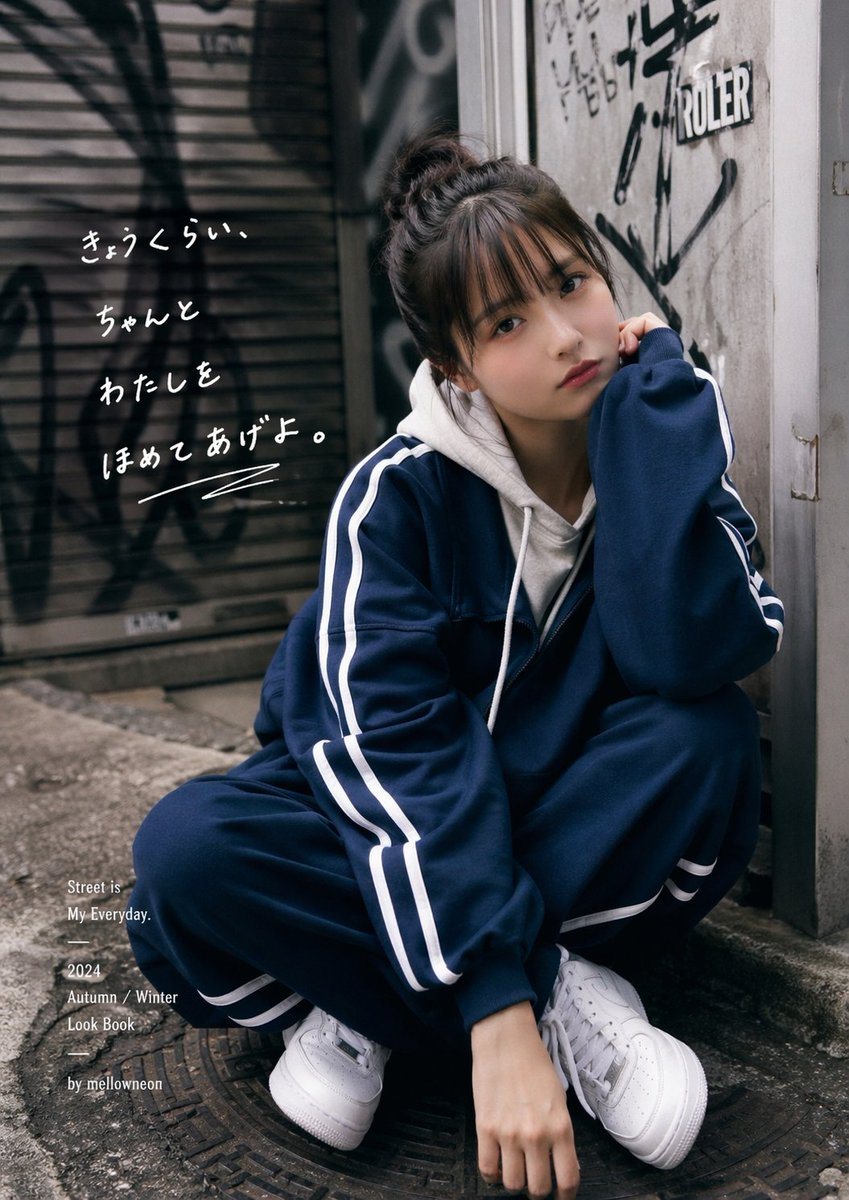 A moody urban fashion editorial photo of a young woman crouching in a narrow alley beside a worn concrete wall covered with black graffiti and stickers, with a metal shutter in the backgr...