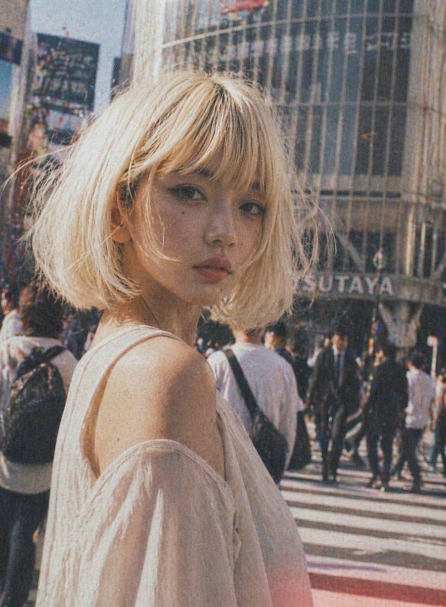A candid street-fashion portrait of a young woman standing at a busy Tokyo crosswalk in daylight, framed from the upper torso up and turned slightly away from the camera so her bare shoul...
