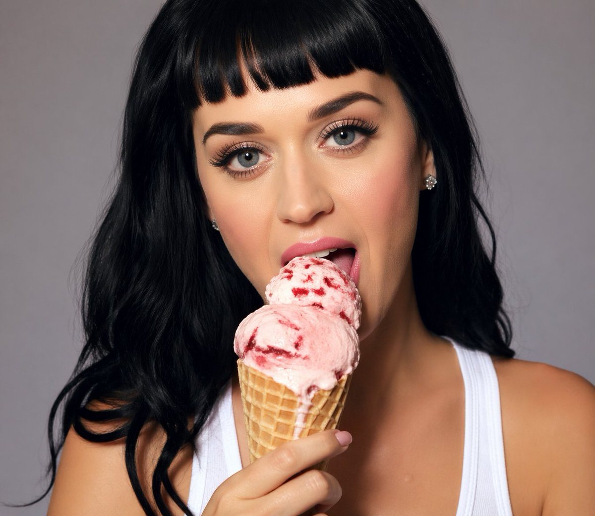 A polished studio portrait of a glamorous pop-star-like woman from the chest up, centered against a smooth neutral gray backdrop. She has jet black long loose wavy hair with a middle part...