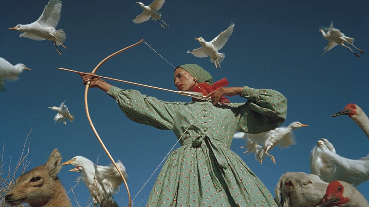 A surreal cinematic outdoor portrait of a masked archer standing in a dry field under a deep clear blue sky, framed from about the knees up in a wide horizontal composition. The central f...