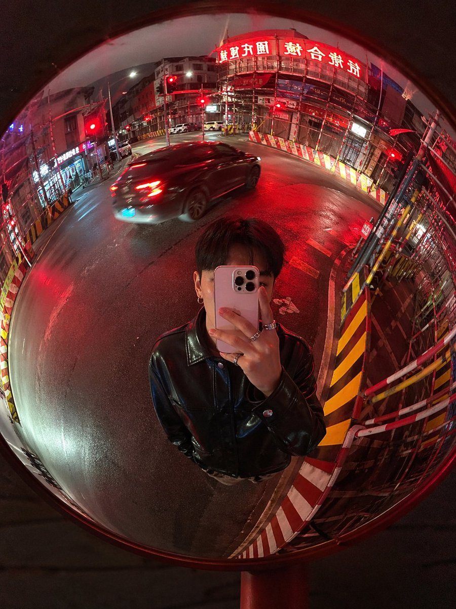 A realistic nighttime street photo shot as a mirror selfie through a large roadside convex traffic mirror, vertical 3:4 composition, with the entire circular mirror filling the frame and...