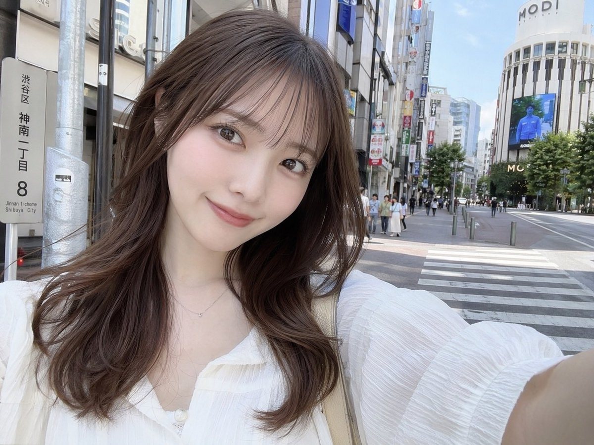 A photorealistic selfie of a young Japanese woman standing on a sunny city street in Shibuya, Japan. She has a soft smile, fair skin, light makeup, and long wavy brown hair with see-throu...
