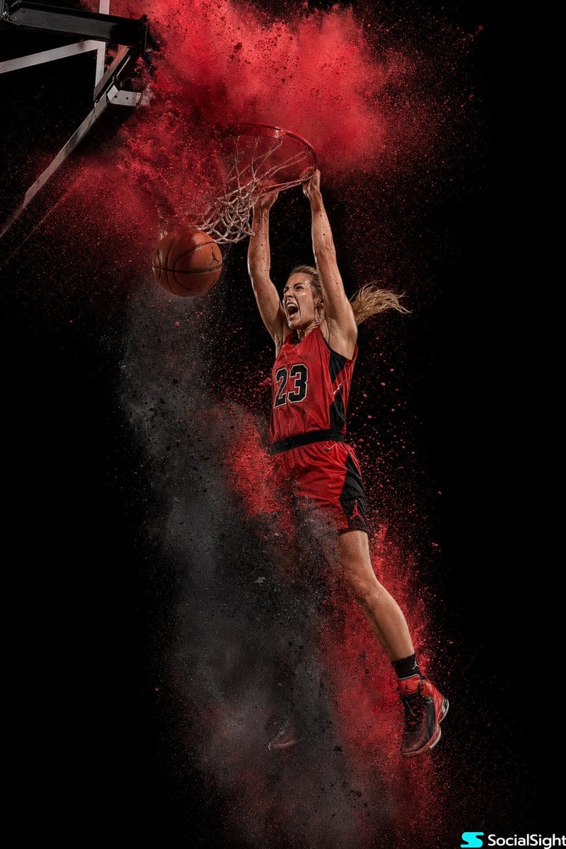 A female basketball player, fully elevated mid-air at the peak of a two-handed dunk, snapping the rim. Her face shows intense focus and exertion. Attire: Classic red and black basketball...