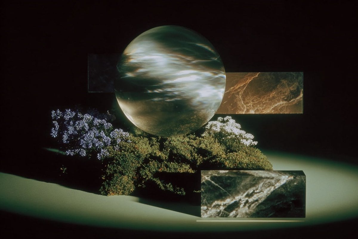 A surreal still-life in a dark black void, staged like a moody luxury sculpture display under a single soft spotlight. At the center, a large floating sphere dominates the composition, ab...