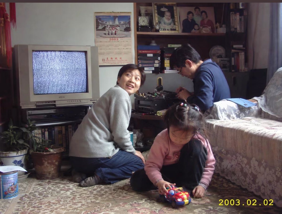 A candid documentary-style indoor family snapshot in a modest early-2000s living room in China, photographed like a real consumer camera photo with soft ambient daylight, slight grain, na...