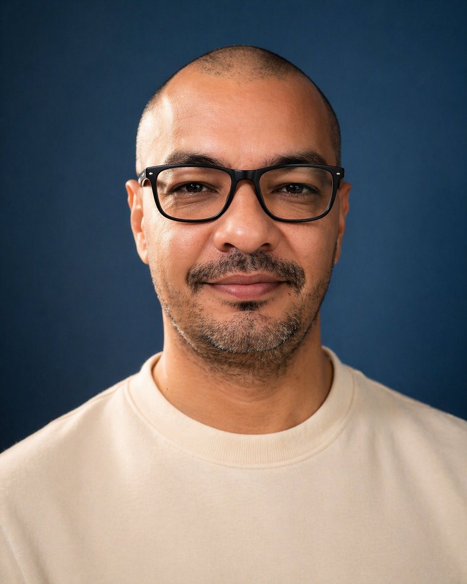 A centered studio portrait photo of a single adult man from upper chest to top of head, facing straight toward the camera against a smooth deep navy-blue backdrop. He has a very closely s...