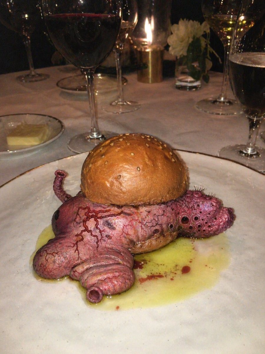 A dark, moody fine-dining restaurant scene photographed with direct flash and a slightly unsettling surreal-horror tone. Center the composition on a plated gourmet sandwich that looks lik...