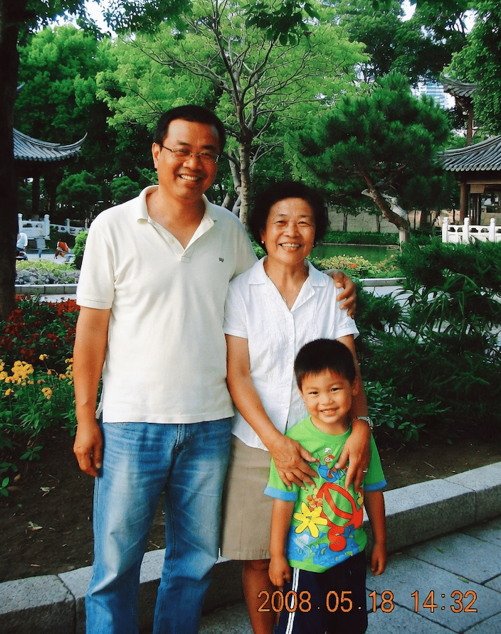 A candid family snapshot styled like a consumer digital camera photo from around 2008 in China, showing a three-person family standing together in a traditional Chinese park garden. Verti...