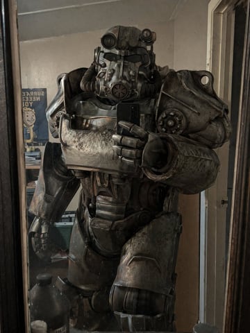 A person wearing a highly detailed, weathered, and battle-damaged T-60 power armor from Fallout taking a mirror selfie with a black smartphone. The armor is bulky, metallic, and covered i...