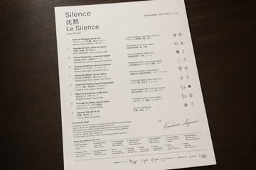 A photorealistic top-down product-style photograph of a single luxury tasting menu printed on bright white textured paper and placed on a dark walnut wooden tabletop, with soft warm ambie...