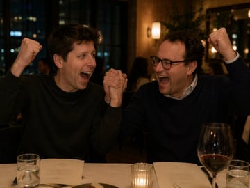 A candid, warm-toned photograph of two men sitting at a table in a dimly lit upscale restaurant at night. On the left is a man with dark hair wearing a dark grey crewneck sweater. On the...