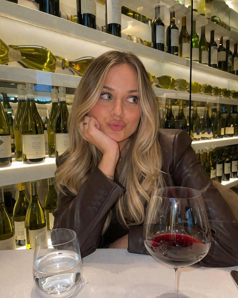 A photorealistic, casual smartphone photo of a young woman with long blonde balayage hair sitting at a table in a wine bar. She is wearing a dark brown leather blazer. She is resting her...