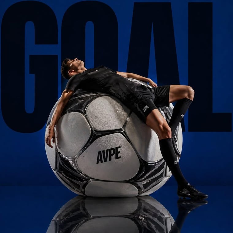 A dramatic sports editorial scene featuring a professional male footballer wearing an all-black kit, reclining confidently on top of an oversized soccer ball. The ball is hyper-detailed w...